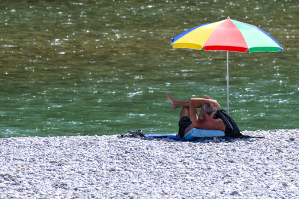 Ein Mann liegt unter einem bunten Sonnenschirm am Ufer der Isar.