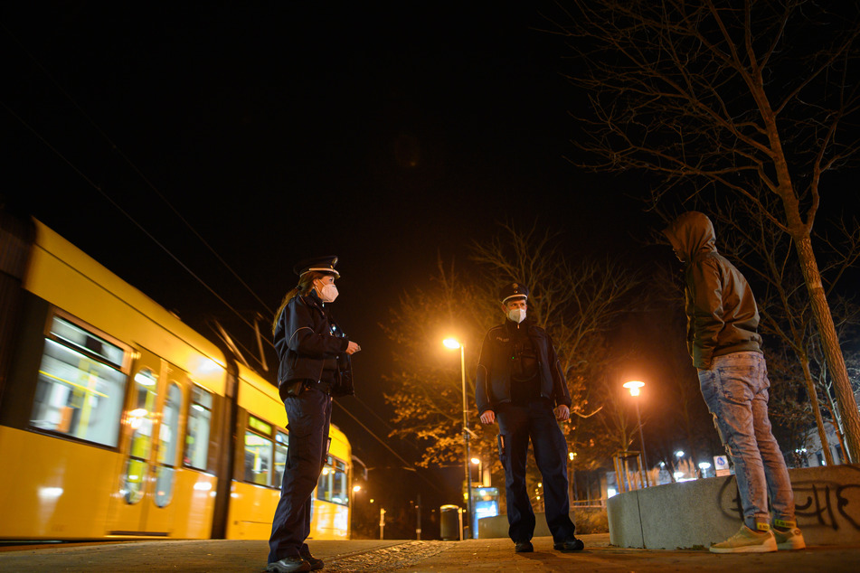 Polizisten überprüfen in der Nacht im Rahmen einer Kontrolle der Ausgangssperre einen Mann an einer Haltestelle. An einigen Orten in NRW dürfen die Menschen nachts nur noch aus wichtigen Gründen vor die Tür (Symbolbild).