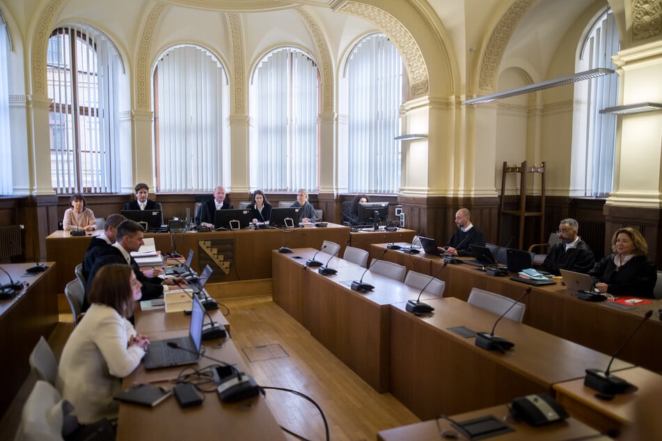 Die Anklagebank bleibt leer, trotzdem beginnt das Gericht die Hauptverhandlung. Die Angeklagten halten sich derweil in einem vom Gericht zur Verfügung gestellten Hinterzimmer versteckt.