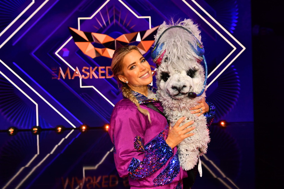 Die Moderatorin hält die Maske der Figur des Alpakas bei "The Masked Singer".