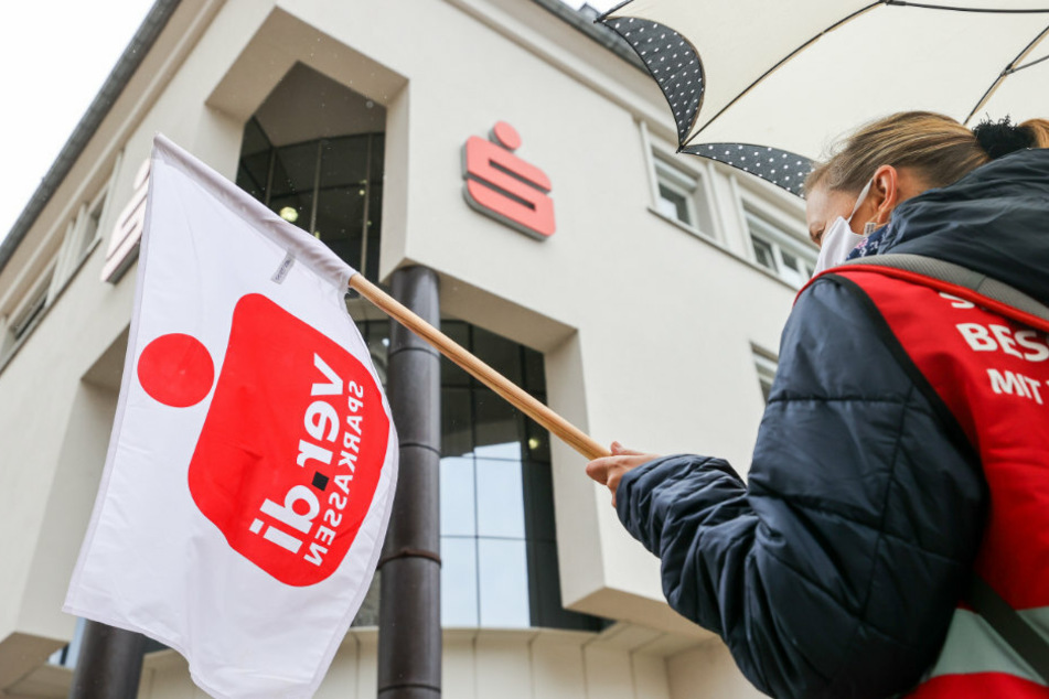 Eine Beschäftigte einer Sparkassen demonstriert mit einer Fahne in der Hand im Rahmen eines Warnstreiks vor der Sparkasse.