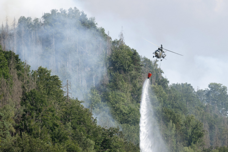 Ein Löschhubschrauber fliegt mit einem Löschwasser-Außenlastbehälter über einen Brand im Nationalpark Sächsische Schweiz.
