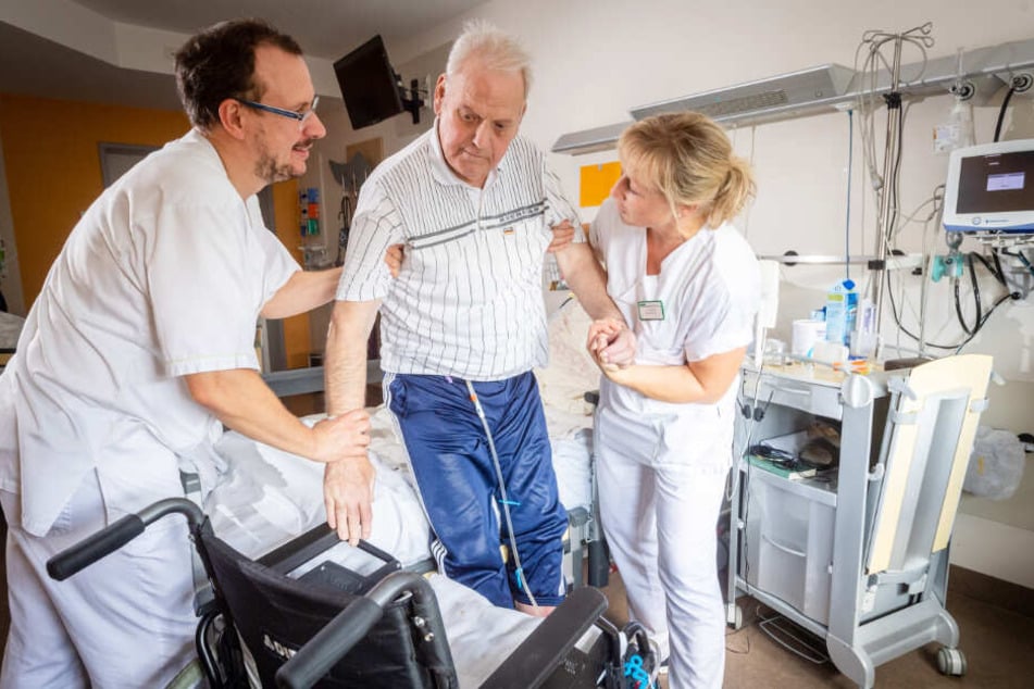 Stationsleiterin Jana Rohatzsch (re.) und Krankenpfleger Frank Degen (li.) helfen einem Patienten dabei, sich in den Rollstuhl zu setzen.