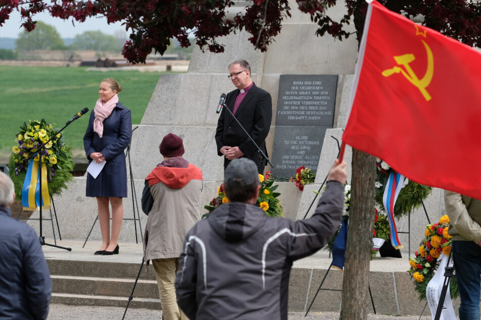 Torgaus Oberbürgermeisterin Romina Barth (CDU) und Johann Schneider, Regionalbischof Halle-Wittenberg der evangelischen Kirche in Mitteldeutschland, stehen am Denkmal der Begegnung, im Vordergrund hält ein Teilnehmer die Fahne der Sowjetunion.