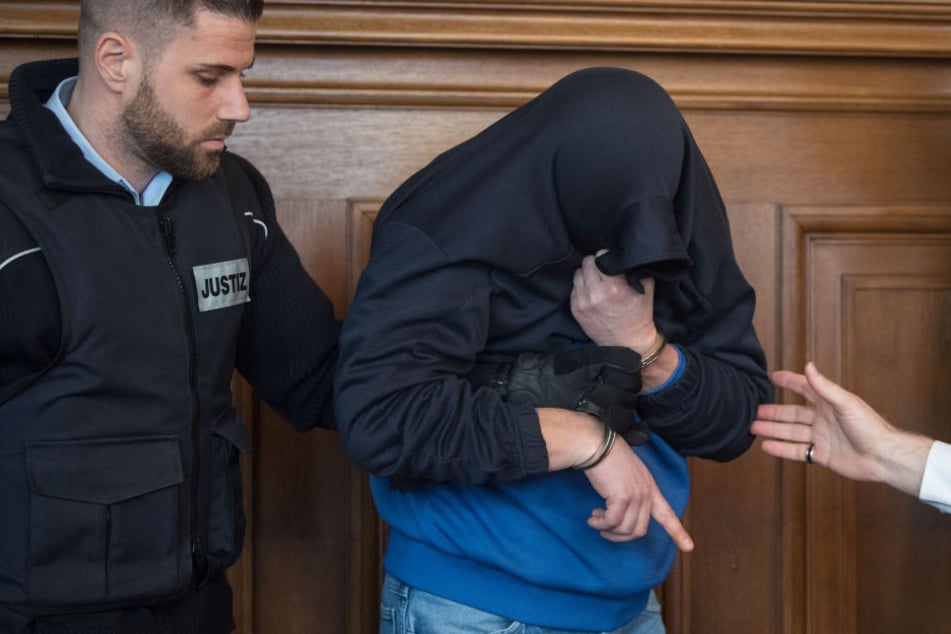 Der Angeklagte im Prozess um einen Dreifach-Mord (M) wird im Landgericht im Gerichtssaal von Justizbeamten an seinen Platz geführt. (Archivbild)