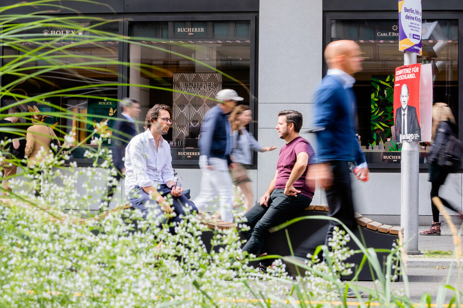 Zwei Männer sitzen auf dem autofreien Teil der Friedrichstraße in Berlin-Mitte vor Schaufenstern, wobei Passanten vorbeigehen.
