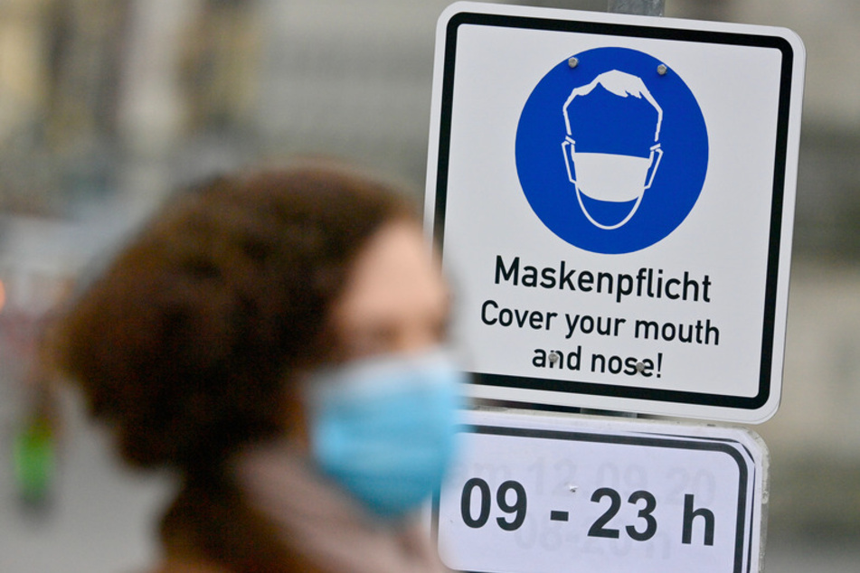 An einem Schild mit der Aufschrift "Maskenpflicht - Cover your mouth and nose!" in einer Fußgängerzone in der Innenstadt geht eine Frau mit Nase-Mund-Bedeckung vorbei.