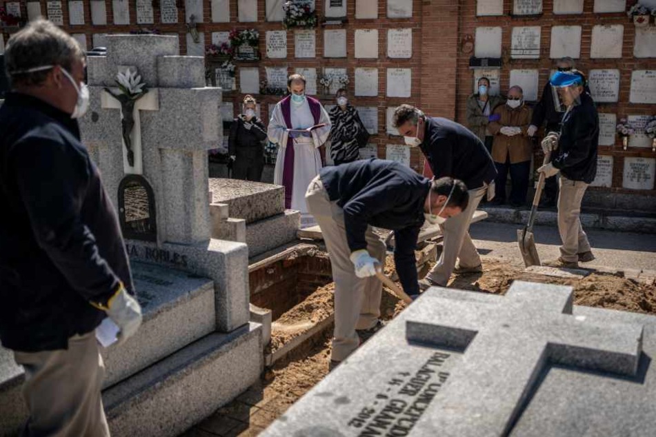Spanien, Madrid (28. März): Während einer Beerdigung eines an dem Coronavirus verstorbenen Menschens beten ein Priester und Familienangehörige hinter Totengräbern, allesamt Mundschutzmasken tragend, am Grab auf dem Friedhof Almudena.