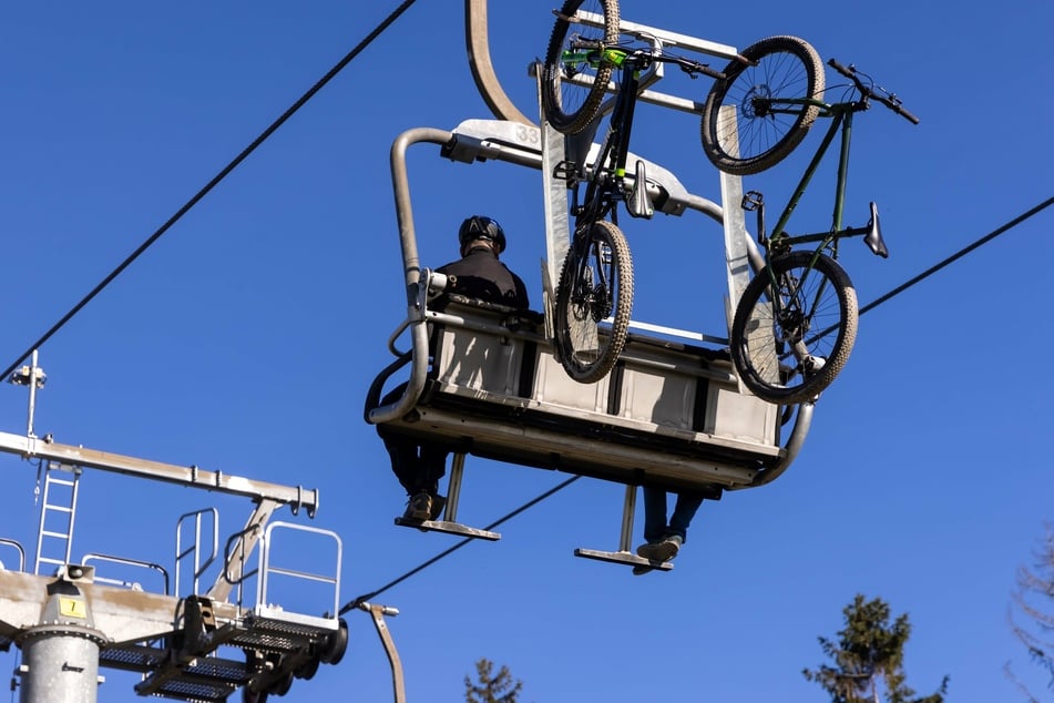 In Schöneck fahren Mountainbiker mit dem Skilift den Berg hoch. Ihre Räder hängen hinter den Sitzen.