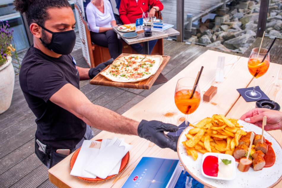 Ein Kellner bringt Gästen mit Maske ihr bestelltes Essen an den Tisch.