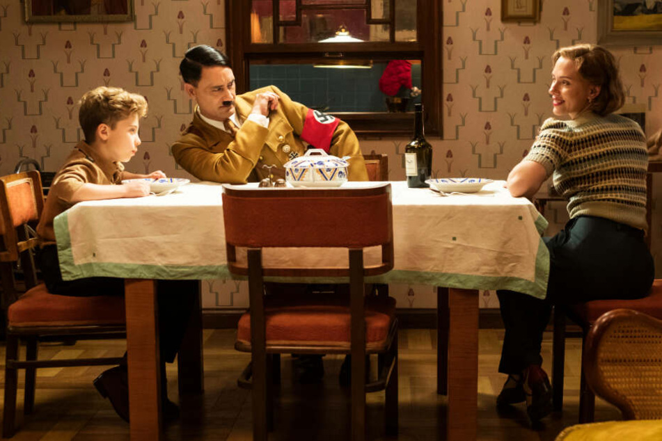 Jojo Rabbit (l., Roman Griffin Davis) hat seinen unsichtbaren Freund Adolf Hitler (M., Taika Waititi) immer dabei - auch beim Essen mit seiner Mutter Rosie (Scarlett Johansson).