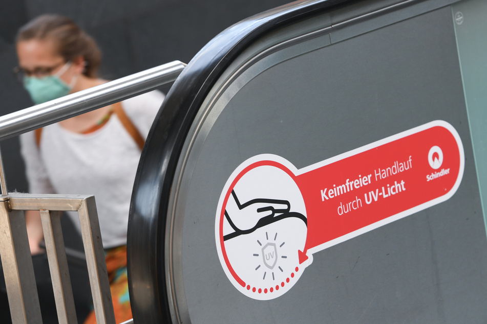 Eine Frau, die einen Mundschutz trägt, geht im Frankfurter Hauptbahnhof an einer Rolltreppe vorbei, die mit einem Aufkleber mit der Aufschrift "Keimfreier Handlauf durch UV-Licht" gekennzeichnet ist. Die Deutsche Bahn will in Zeiten der Corona-Pandemie noch mehr für Hygiene und Sauberkeit in Zügen und an den Bahnhöfen tun.