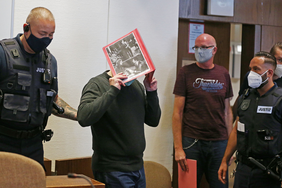 Der Angeklagte hält sich eine Mappe mit aufgeklebten Fotos von Filmszenen des Films "Der Strafverteidiger" vor das Gesicht.