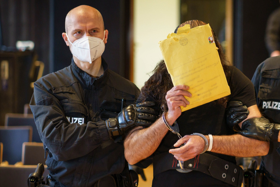 Der 46-jährige Angeklagte wird von Polizisten in Handschellen zur Anklagebank geführt.