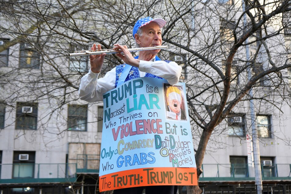An anti-Donald Trump protester plays patriotic tunes on a flute while wearing a sign that shares critical sentiments about the former president.