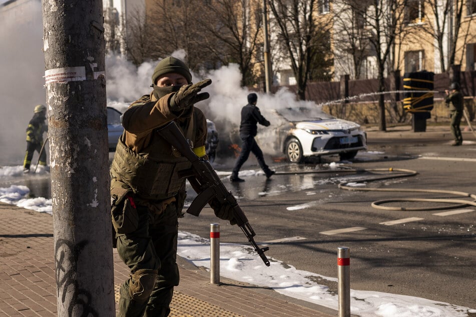 Ukrainische Soldaten beziehen Stellung vor einer Militäreinrichtung, während Feuerwehrleute zwei brennende Autos löschen.