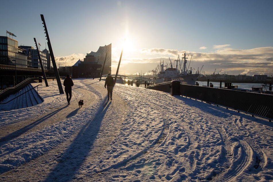 Passanten mit Hund sind auf der verschneiten Elbpromenade bei strahlendem Sonnenschein an den Landungsbrücken im Hafen unterwegs.
