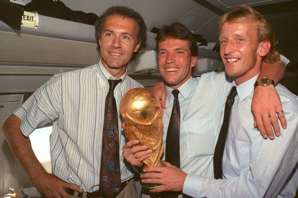 Im Flugzeug 1990 präsentieren der damalige DFB-Teamchef Franz Beckenbauer (l.), Kapitän und Mittelfeldspieler Lothar Matthäus (M.) und Abwehrspieler Andreas Brehme den gewonnenen WM-Pokal.