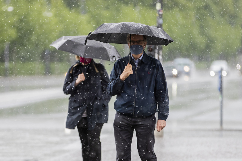 Ein Mann und eine Frau gehen bei starkem Regen mit Mund-Nasen-Schutz und Regenschirmen über den Platz des 18. März in Berlin.