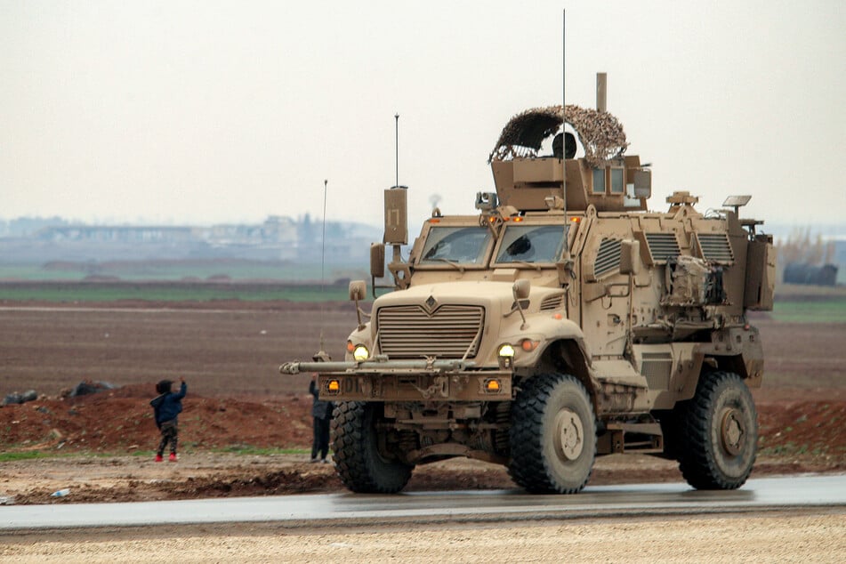 Boys along a road wave to an approaching US military mine-resistant ambush protected (MRAP) armored fighting vehicle moving in a convoy transporting Islamic State group detainees being transferred to Iraq from Syria, on the outskirts of Qahtaniyah in Syria's northeastern Hasakah province on February 7, 2026.
