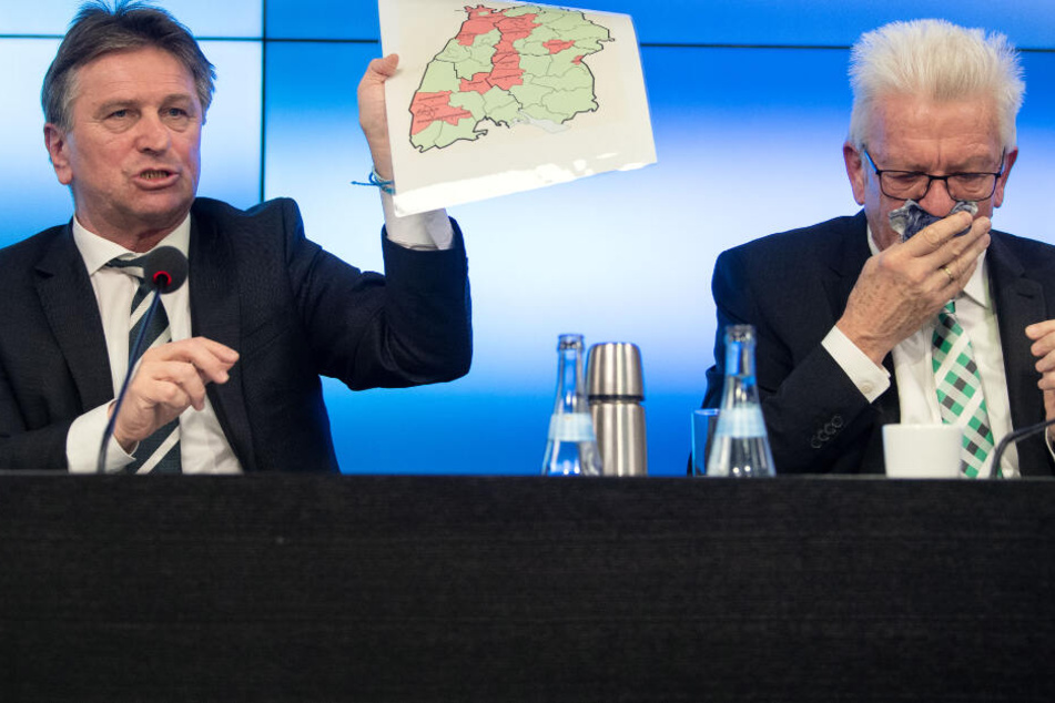 Gesundheitsminister Manfred Lucha (Grüne, l.), hält eine Karte von Baden-Württemberg in der Hand, auf der die Anzahl der Coronavirus-Fälle im Land verzeichnet ist. Ministerpräsident Winfried Kretschmann (Grüne) hält sich ein Taschentuch vor.