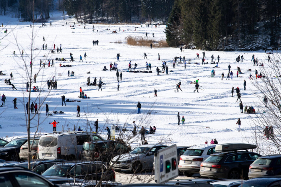Parkende Autos von Ausflüglern sind vor dem zugefrorenen Spitzingsee zu sehen, auf dem sich zahlreiche Touristen und Einheimische tummeln.