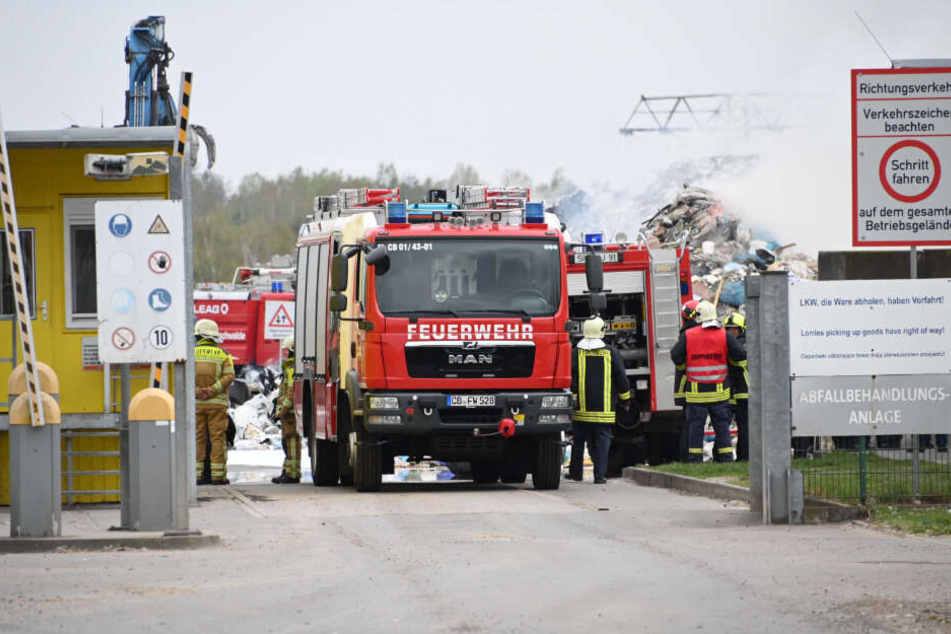 Feuerwehrleute stehen auf einem Recyclinghof vor einem qualmenden Müllberg.