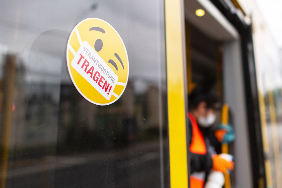 Ein Smiley-Aufkleber mit einem Mundschutz und der Aufschrift "Verantwortung tragen" klebt an der Tür einer Straßenbahn, während eine Mitarbeiterin der Dresdner Verkehrsbetriebe die Handläufe desinfiziert.