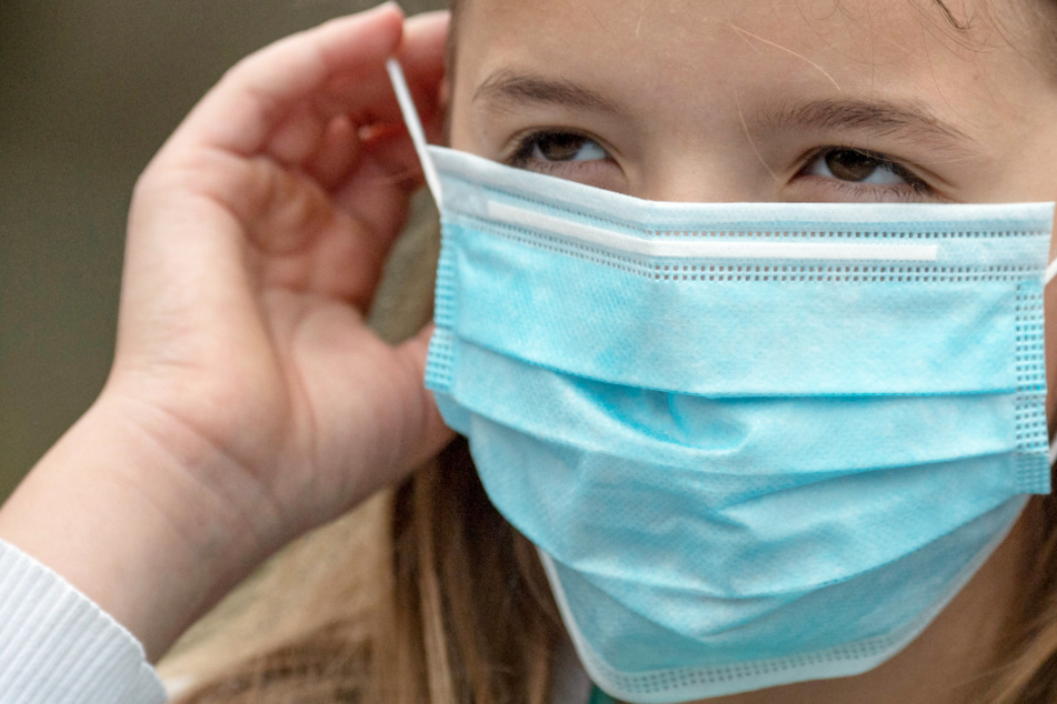 Eine Grundschülerin setzt sich vor dem Betreten eines Schulgeländes eine Einweg-Maske auf.