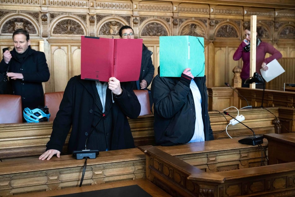 Zwei der Angeklagten stehen vor Prozessbeginn im Gerichtssaal und verbergen ihre Gesichter.