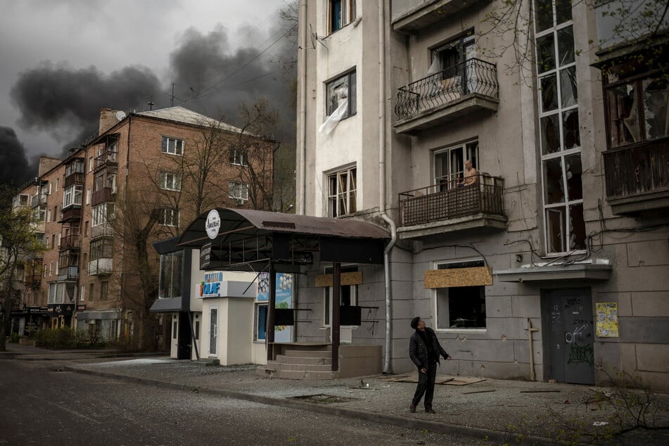 A man stands outside a damaged apartment block in Kharkiv as smoke rises in the background following Russian shelling.