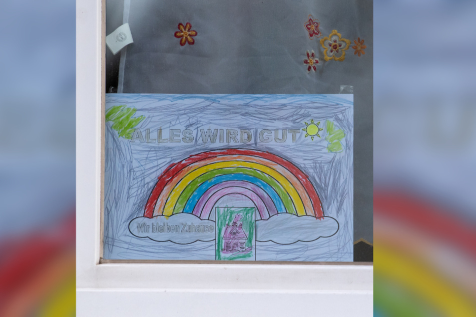 Ein Blatt Papier mit einem Regenbogen und der Aufschrift "Alles wird Gut - Wir bleiben Zuhause" hängt in einem Fenster zur Strassenseite in München.