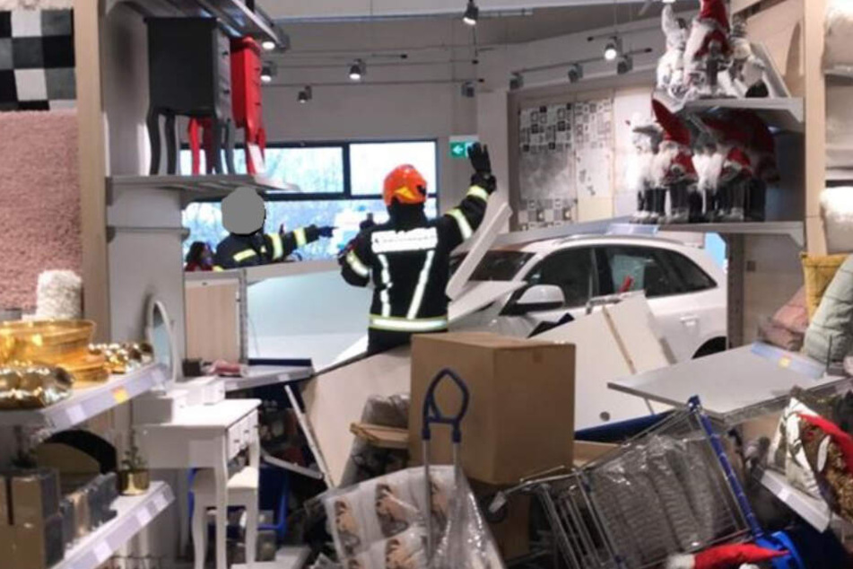 Einsatzkräfte der Feuerwehr stehen vor einem Auto im Inneren einer Filiale eines Einrichtungshauses zwischen Trümmern, nachdem der Wagen in den Laden gekracht und bis zum Kassenbereich gerollt war.