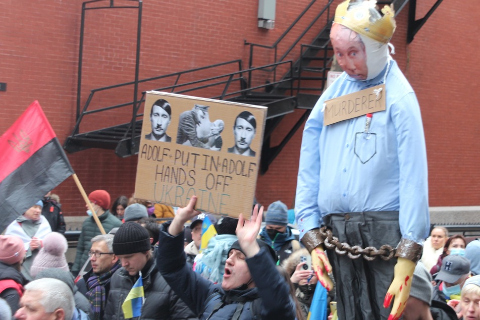 One protester holds a sign that compares Russian president Vladimir Putin to Adolf Hitler. Another protester holds up a life-sized doll with Putin's face, bloody hands, and the sign "MURDERER".