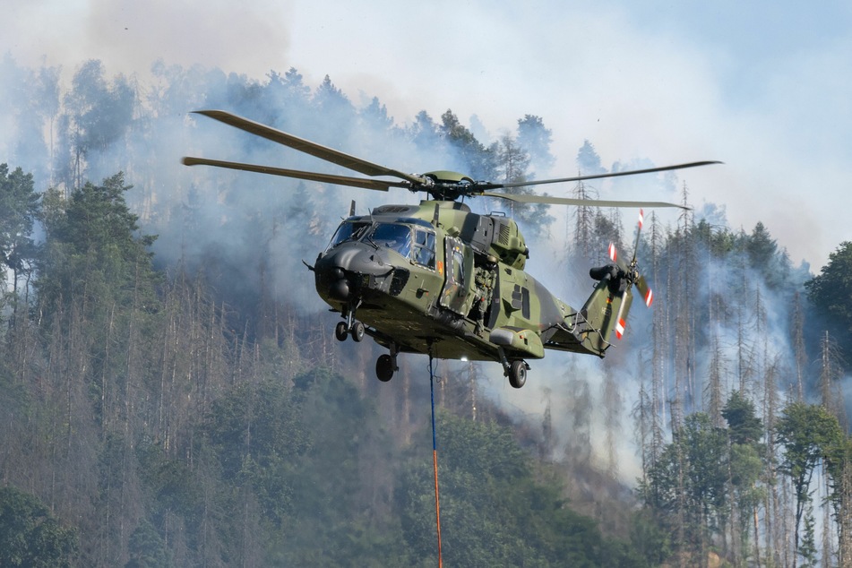 Ein Hubschrauber der Bundeswehr fliegt mit einem Löschwasser-Außenlastbehälter über einen Brand im Nationalpark Sächsische Schweiz.