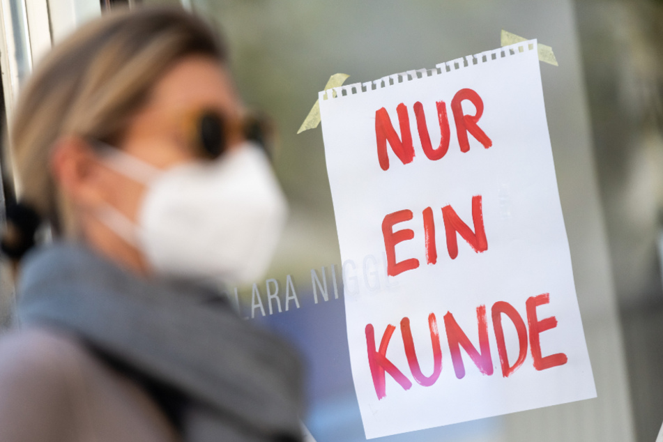 Eine Frau mit Nase-Mund-Schutzmaske geht an einem Geschäft vorbei, an dem ein Schild mit der Aufschrift "Nur ein Kunde" angebracht ist.