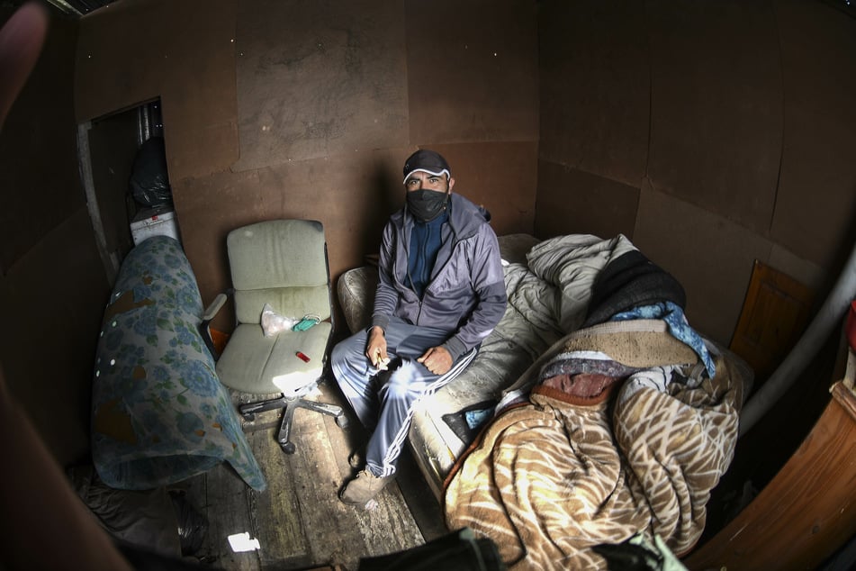 Ein Mann mit Mundschutz sitzt inmitten der Corona-Pandemie auf einer Matratze in einer Hütte aus Pappe in einem bestzten Landstück.
