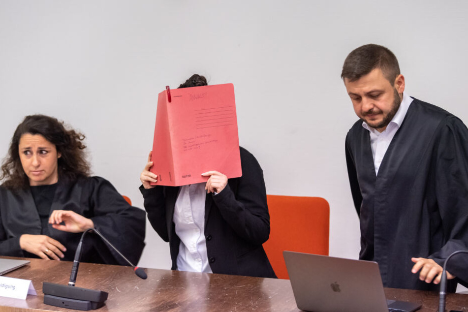 Die Angeklagte hält sich vor Verhandlungsbeginn im Gerichtssaals einen roten Aktendeckel vors Gesicht. Neben ihr sind ihre Anwälte Sera Basay-Yildiz (l) und Ali Aydin (r).