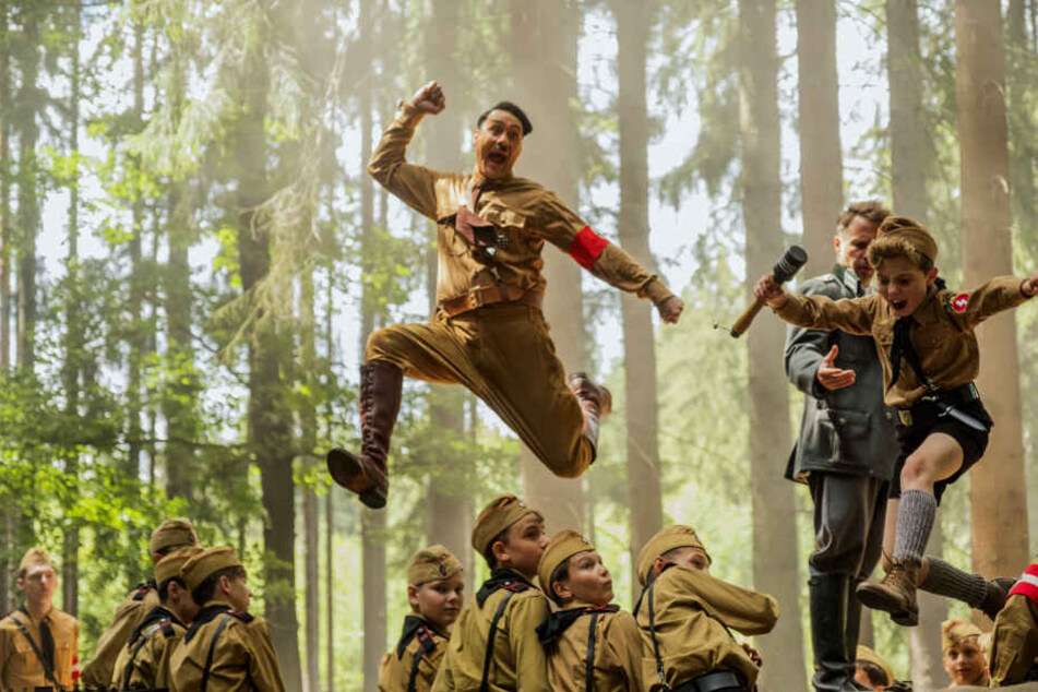 Johannes "Jojo" Betzler (r., Roman Griffin Davis) klaut Captain Klenzendorf (Zweiter von rechts, Sam Rockwell) angestachelt von Adolf Hitler (oben, Taika Waititi) eine Granate aus der Hand.