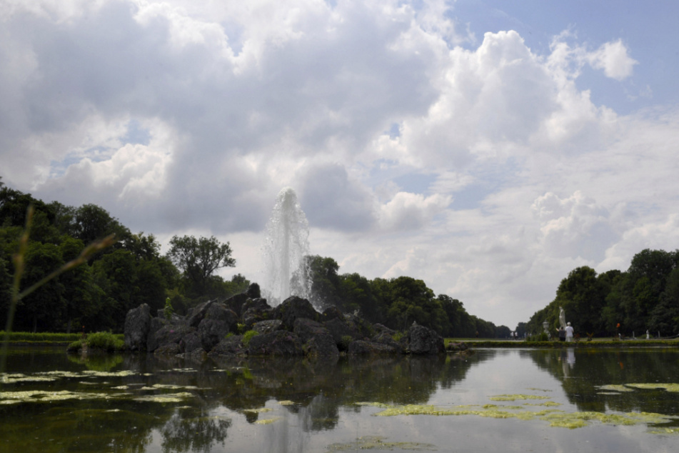 Die Fontäne eines Springbrunnens ist im Schlosspark Nymphenburg bei bewölktem Himmel und Sonne zu sehen.