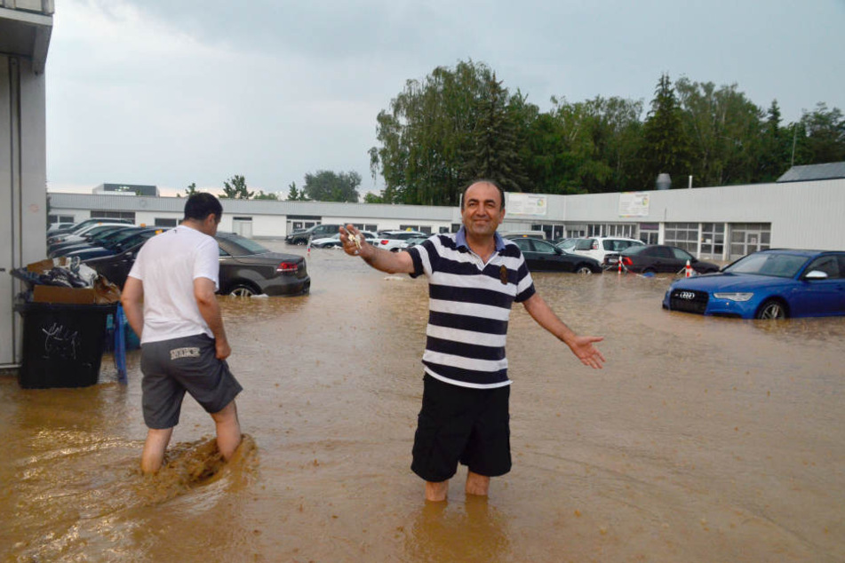 Nach einem schweren Unwetter steht Atmet Acer auf dem Geländer einer KFZ-Reparaturwerkstatt und zeigt seinen Autoschlüssel. Er freut sich, weil er seinen BMW 7er noch aus dem hohen Wasser herausfahren konnte.