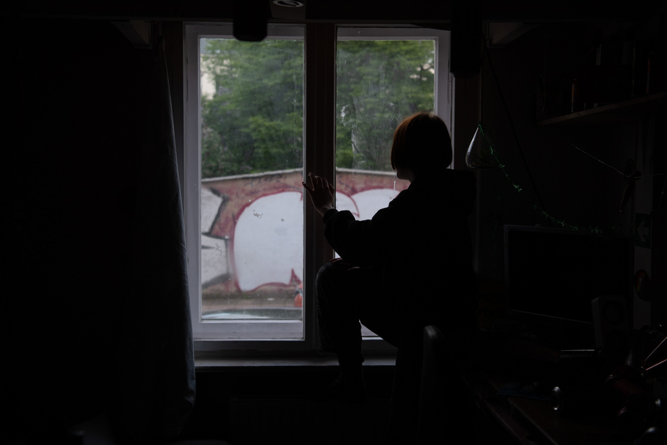 Eine Jugendliche sitzt in einem abgedunkelten Zimmer am Fenster.
