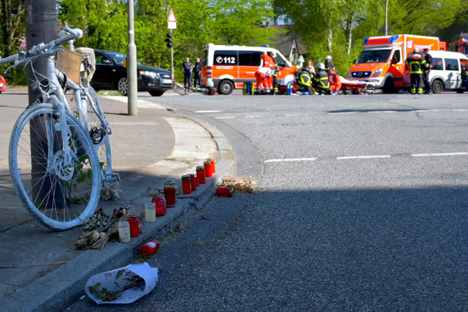 Die kleine Gedenkstätte nach einem tödlichen Radunfall ist im Vordergrund zu sehen, während auf der Unfallkreuzung ein Notarzt einen weiteren verunfallten Fahrradfahrer versorgt.