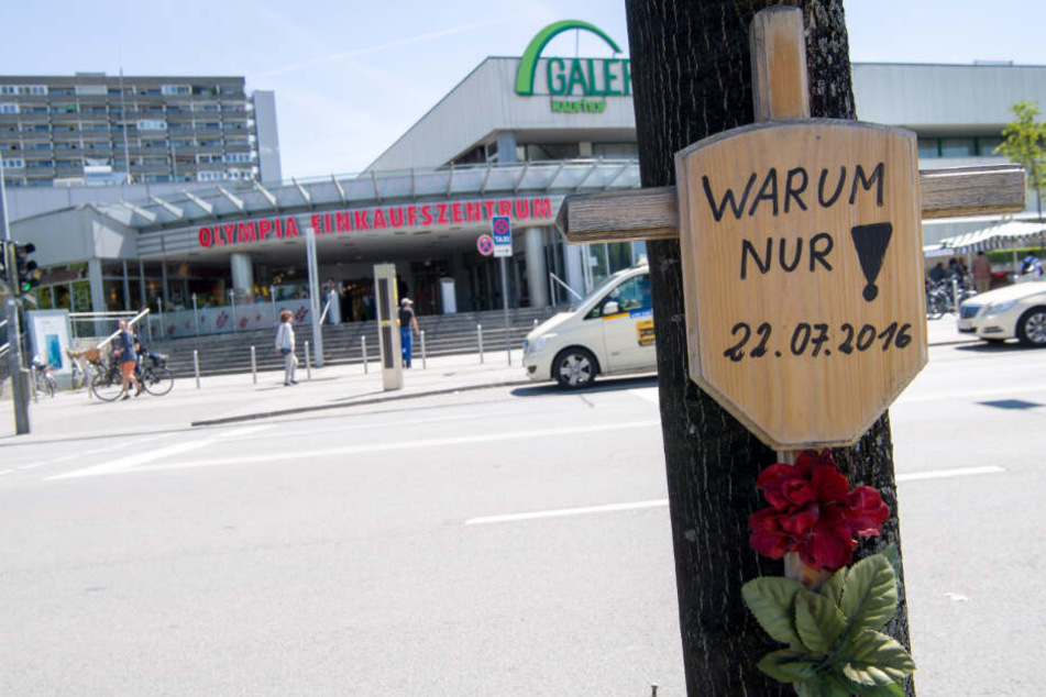Ein Holzkreuz mit einem Schild und der Aufschrift "Warum nur! - 22.07.2016" hängt vor dem Eingang zum Olympia-Einkaufszentrum (OEZ). (Archiv)
