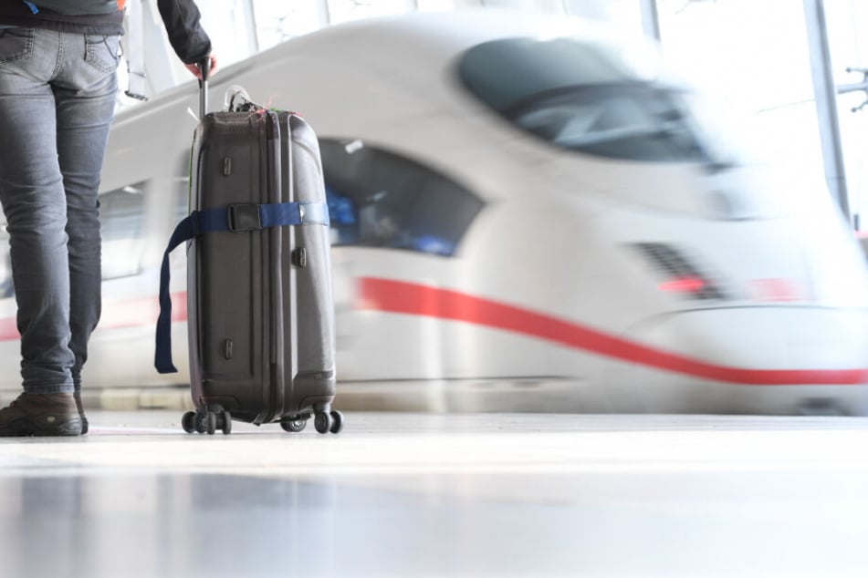 Ein Mann geht mit einem Rollkoffer im Bahnhof an einem losfahrenden ICE vorbei. (Symbolbild)