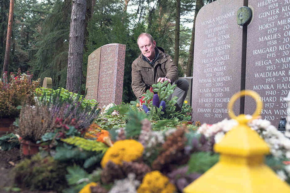 Heidefriedhof-Chef Mario Fischer (50) kniet vor einer Urnen-Gemeinschaftsanlage. Auf der grünen Wiese „mit Namensnennung“ teilen sich 20 Verstorbene ein Grabmal. Ihre Angehörigen platzieren Engel, Fotos und Kerzen.