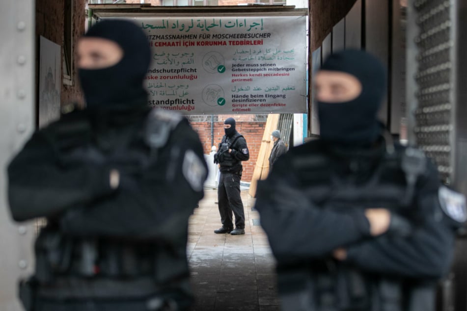 Im Vordergrund stehen zwei Polizisten am Eingang zur Moschee. Im Hintergrund ist ein Plakat zu erkennen, welches die Besucher in drei Sprachen über die geltenden Schutzmaßnahmen informiert.