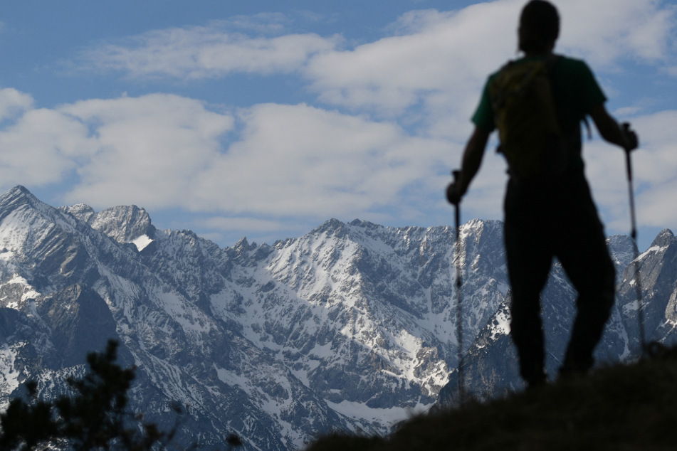 Ein Wanderer steht vor den schneebedeckten Gipfeln des Wettersteingebirges, links ist das Dreieck der Alpspitze und rechts die Zugspitze zu sehen.