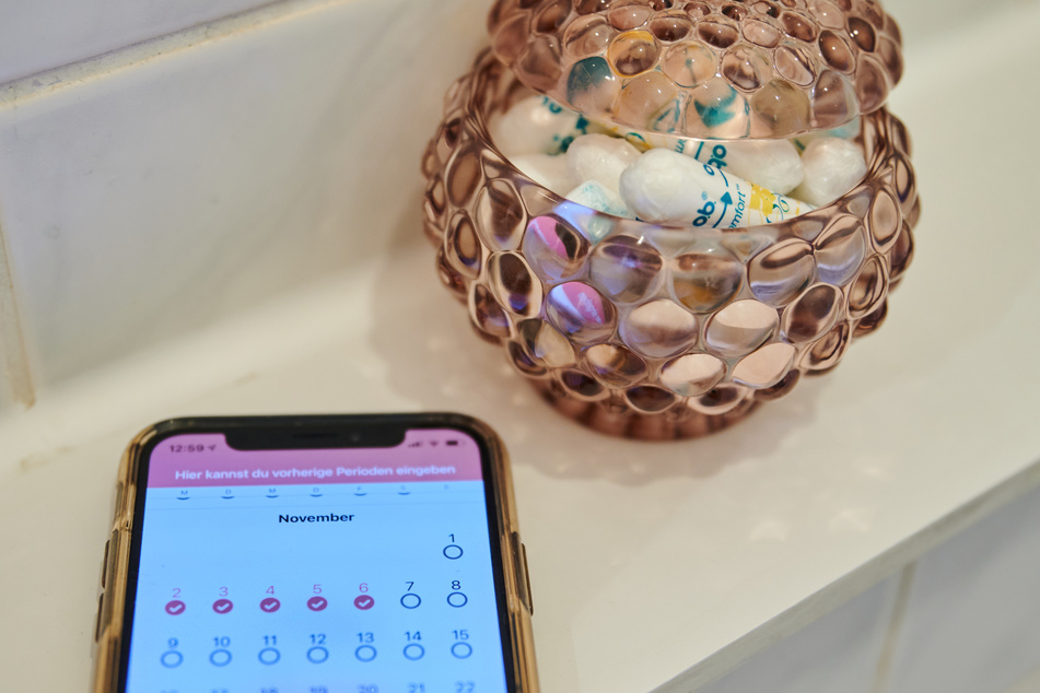 Ein Menstruationskalender auf einem Smartphone liegt im Badezimmer neben einem Behälter mit Tampons.