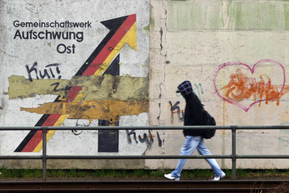 Ein Passant geht an einer Schallschutzmauer an einem aus der Wendezeit stammenden verwitterten Wandbild mit dem Schriftzug "Gemeinschaftswerk Aufschwung Ost" in Magdeburg vorbei.